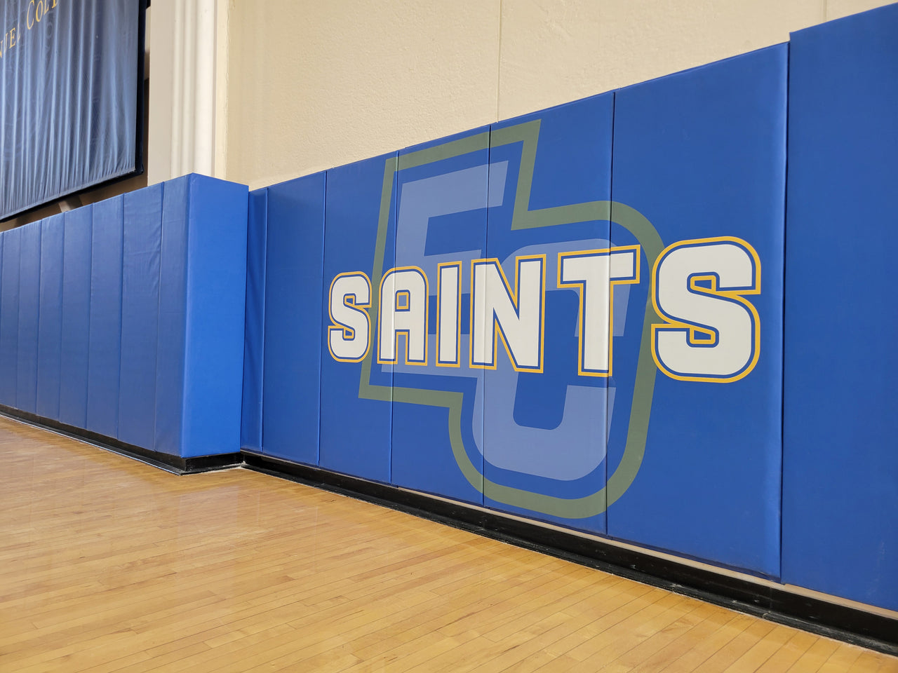 Blue gym wall padding printed with white Saints lettering over a large monogram along a wood gym floor