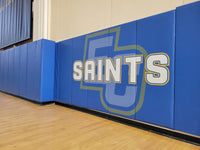 Thumbnail for Blue gym wall padding printed with white Saints lettering over a large monogram along a wood gym floor