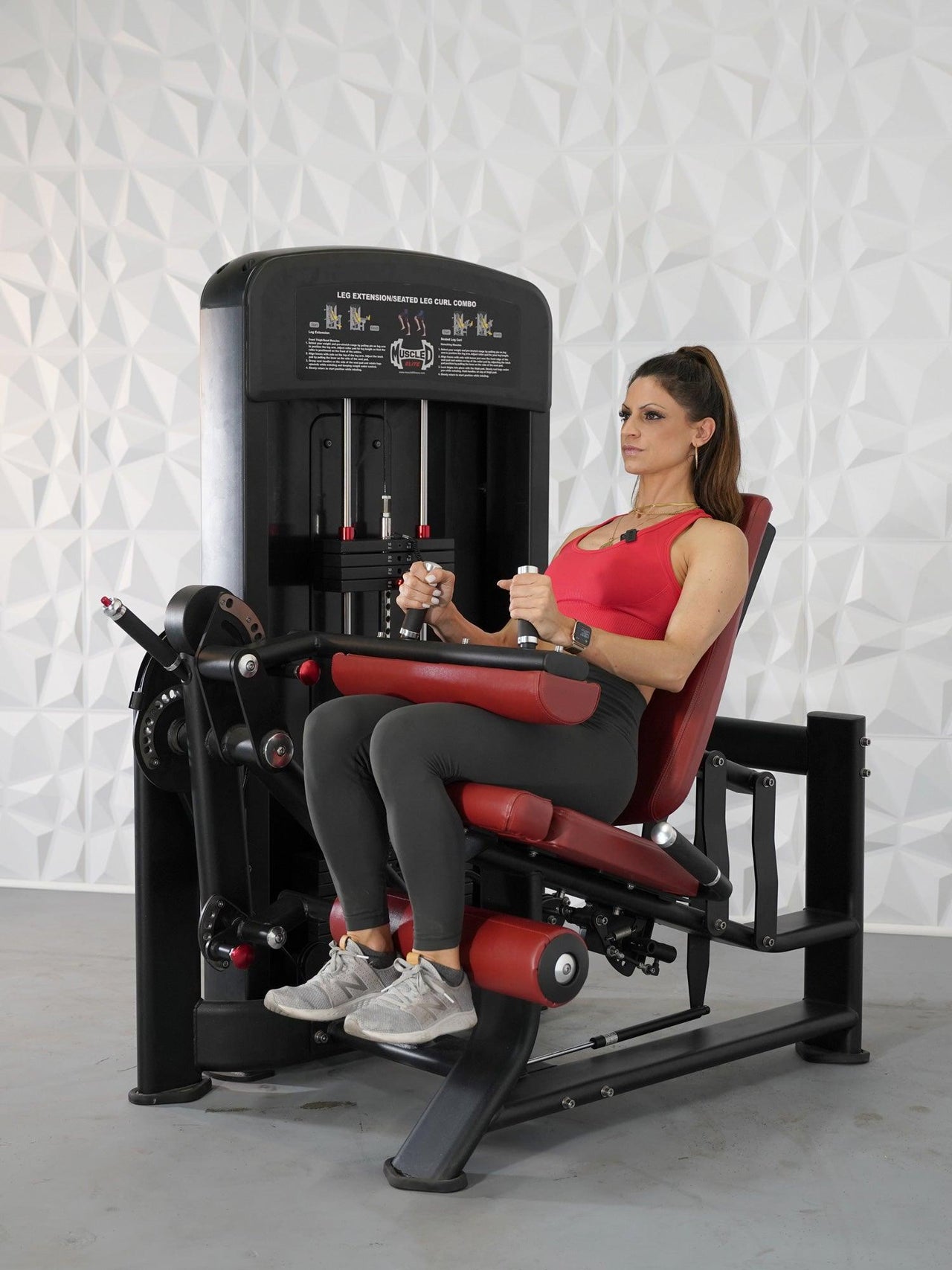 Woman in red top and gray leggings using a black and red leg extension/curl combo machine.