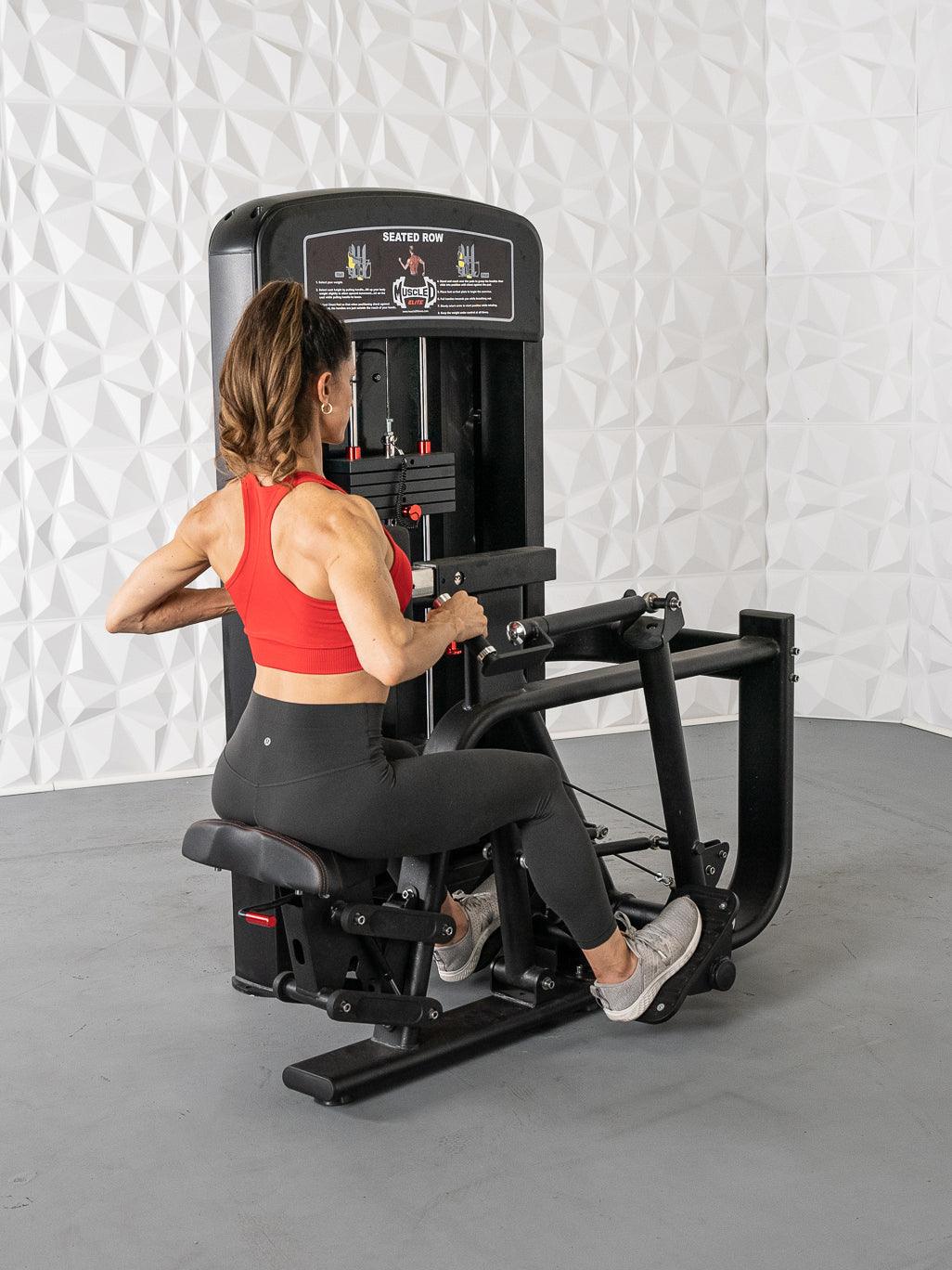 Woman in red top and black leggings using a black seated row machine with white textured wall background.