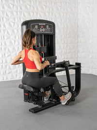 Thumbnail for Woman in red top and black leggings using a black seated row machine with white textured wall background.