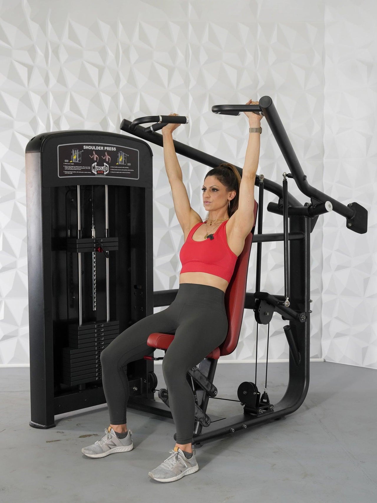 Woman in red top and gray leggings using a black shoulder press machine with red seat.