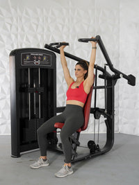 Thumbnail for Woman in red top and gray leggings using a black shoulder press machine with red seat.
