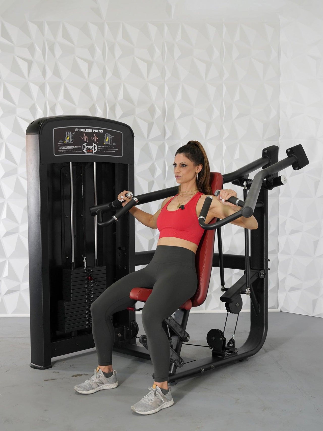 Woman in red top and gray leggings seated on a black and red shoulder press machine.