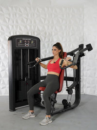 Thumbnail for Woman in red top and gray leggings seated on a black and red shoulder press machine.