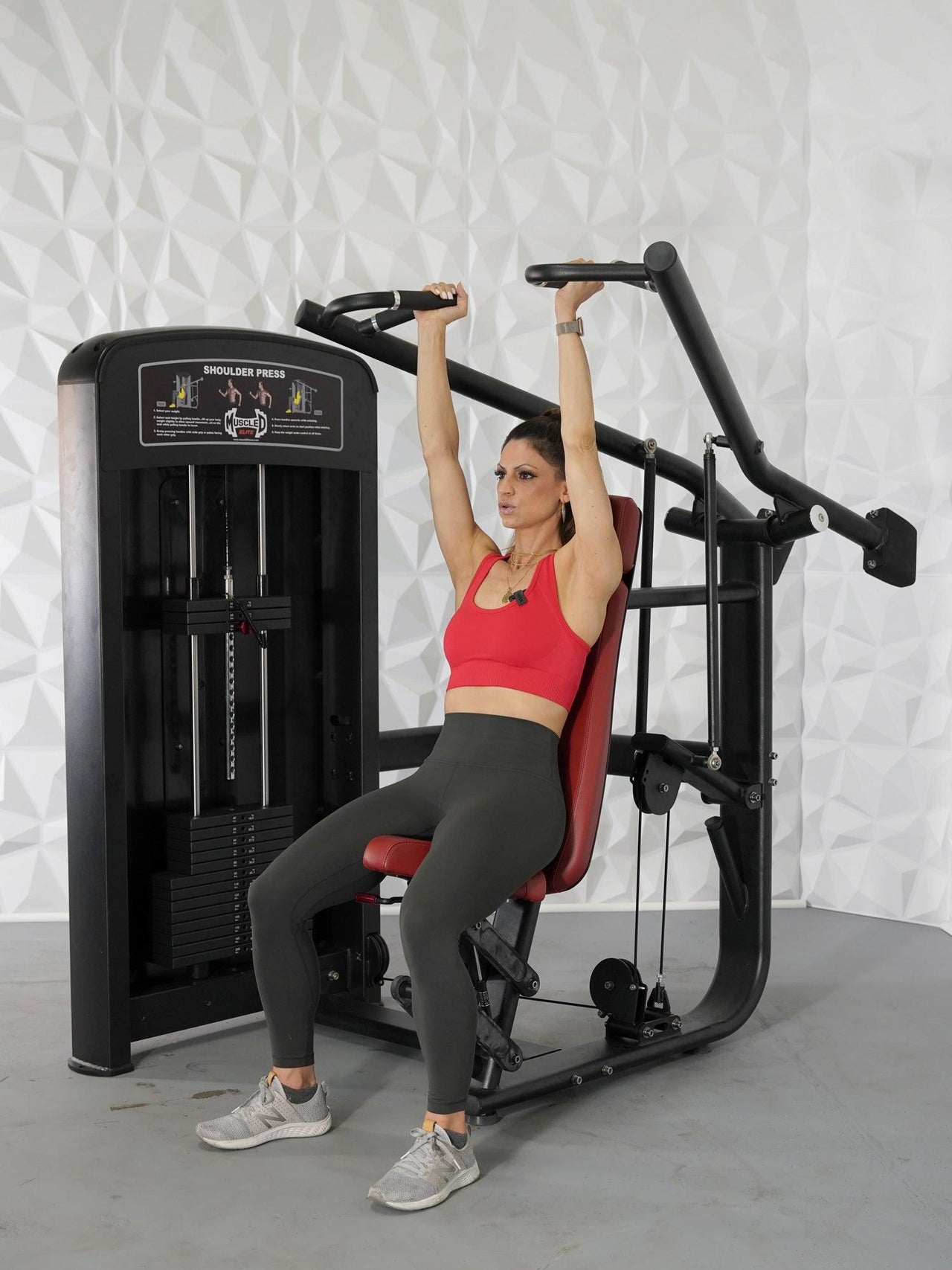 Woman in red top and gray leggings using a black and red seated shoulder press machine.