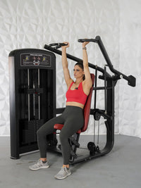 Thumbnail for Woman in red top and gray leggings using a black and red seated shoulder press machine.