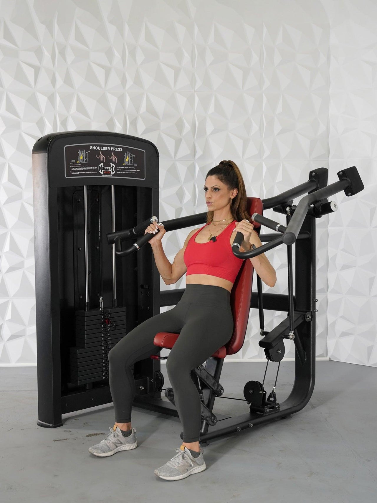 Woman in red top and gray leggings using a black and red shoulder press machine.