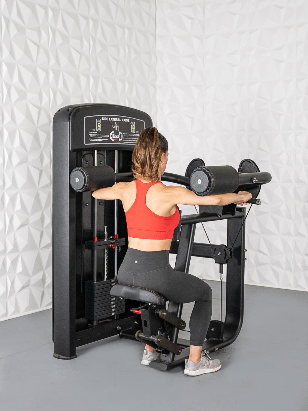 Woman in red top and black leggings using a black side lateral raise machine with padded shoulder rests.