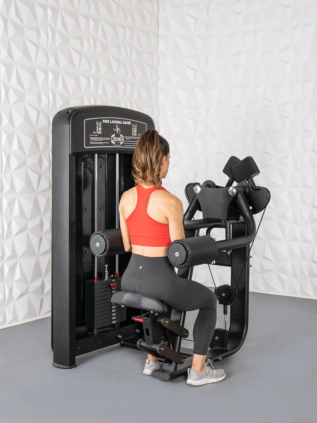 Woman in red top and black leggings using a black side lateral raise machine.