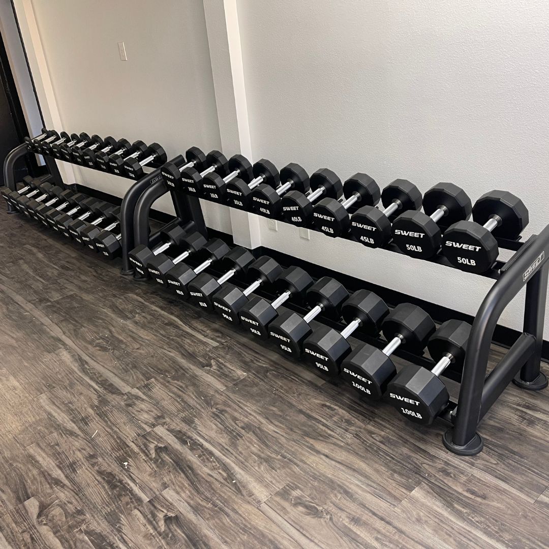 Two black 2-tier dumbbell racks with hexagonal dumbbells in a gym.