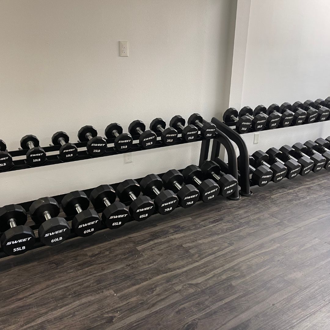 Two black 2-tier dumbbell racks filled with black hex dumbbells on a dark wood floor.