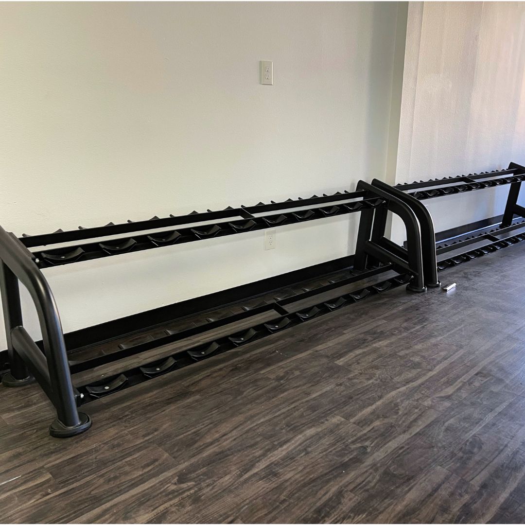 Two empty black 2-tier dumbbell racks on a dark wood floor against a white wall.