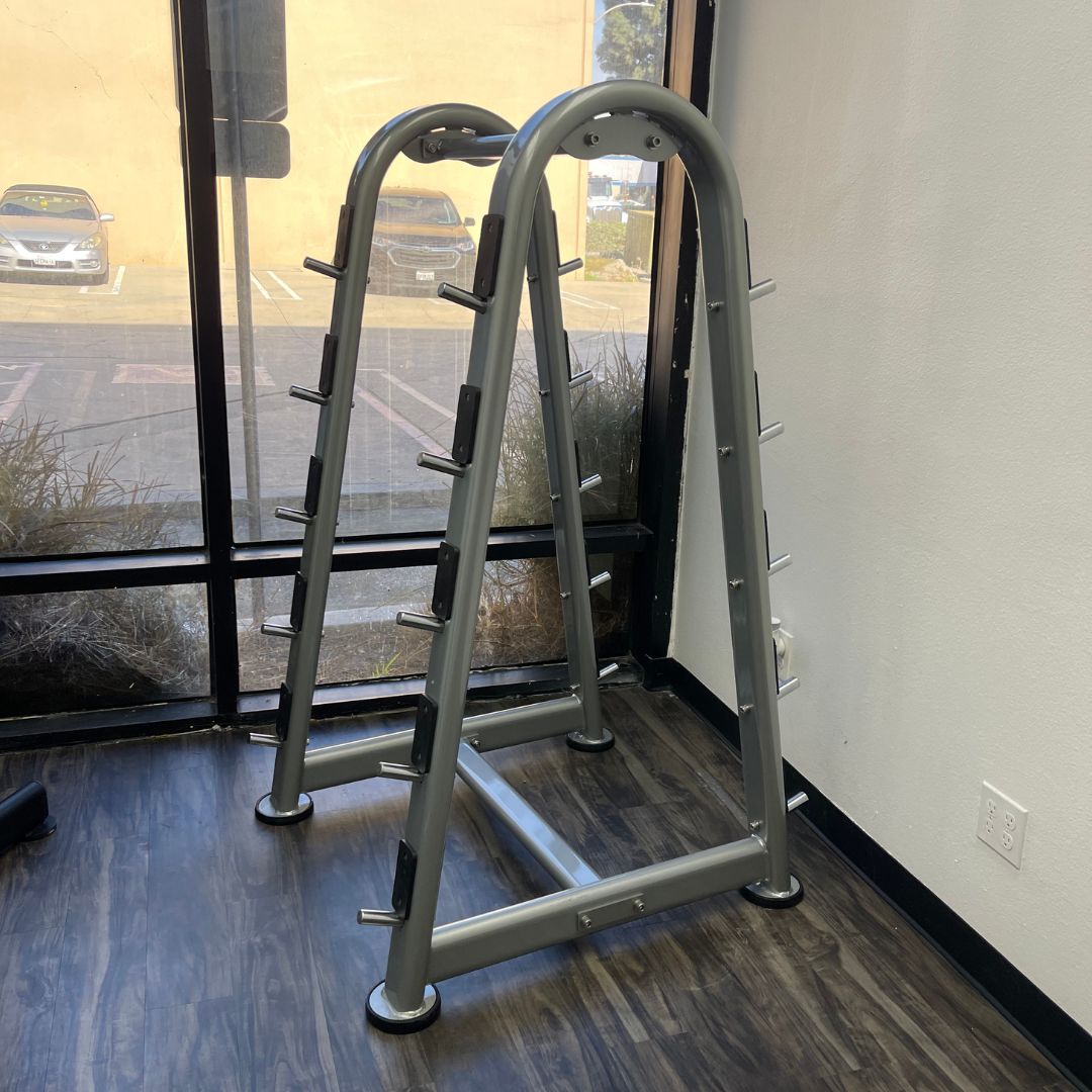 Two gray barbell racks with multiple pegs for weight storage, on a dark wood floor.