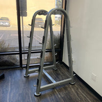 Thumbnail for Two gray barbell racks with multiple pegs for weight storage, on a dark wood floor.