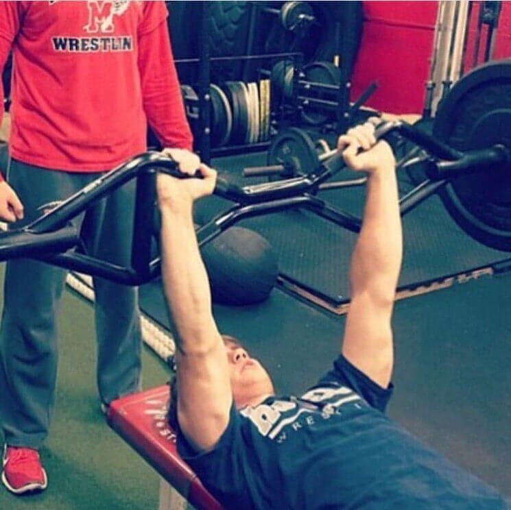 Man bench pressing a multi-grip barbell with a spotter.
