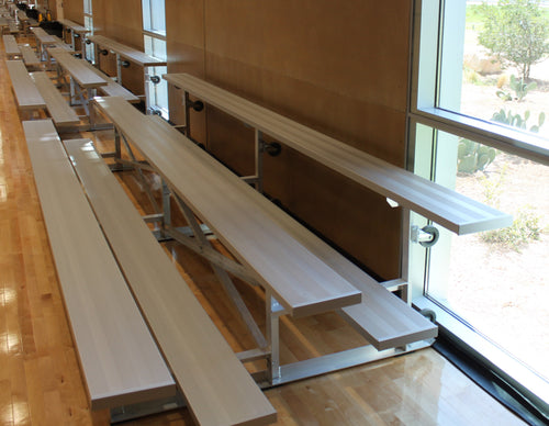 Two-row bleachers with light wood seats and metal frames on a wood floor next to a window.
