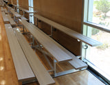 Three rows of light-colored bleachers with metal frames on a wooden floor next to a large window.