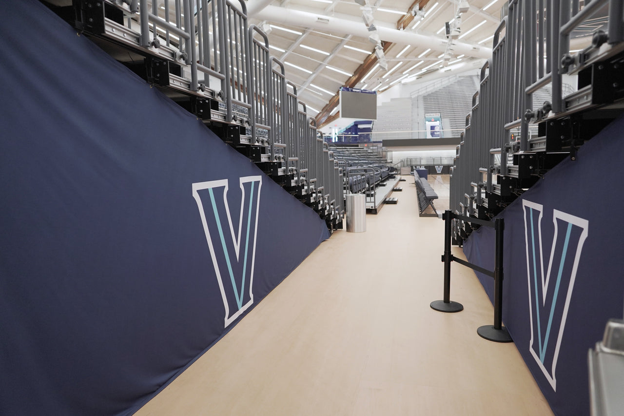 Navy bleacher enclosure covers with white and light blue V logos lining a walkway under arena bleachers