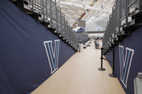 Thumbnail for Navy bleacher enclosure covers with white and light blue V logos lining a walkway under arena bleachers