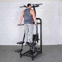 Thumbnail for Man in grey tank top and sweatpants using a black assisted chin/dip machine with white textured wall background.