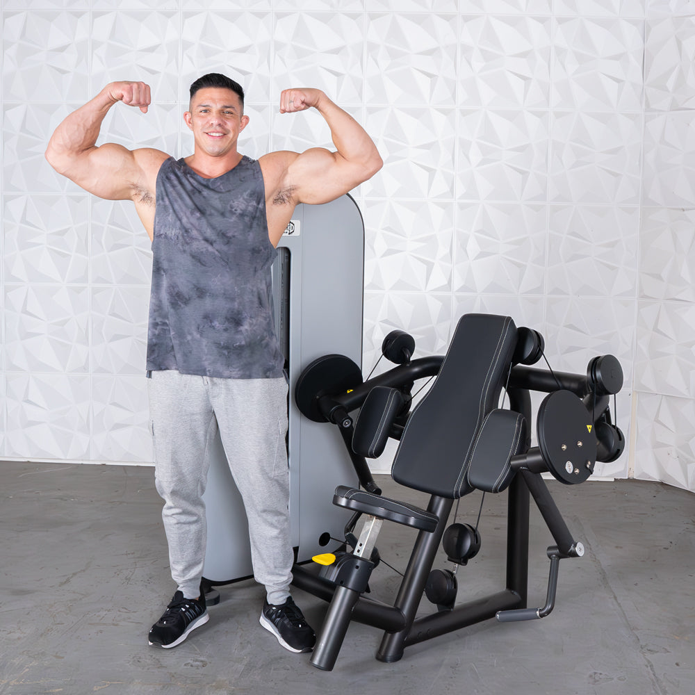 Muscular man flexing next to a black and gray arm curl machine with a padded seat and armrests.