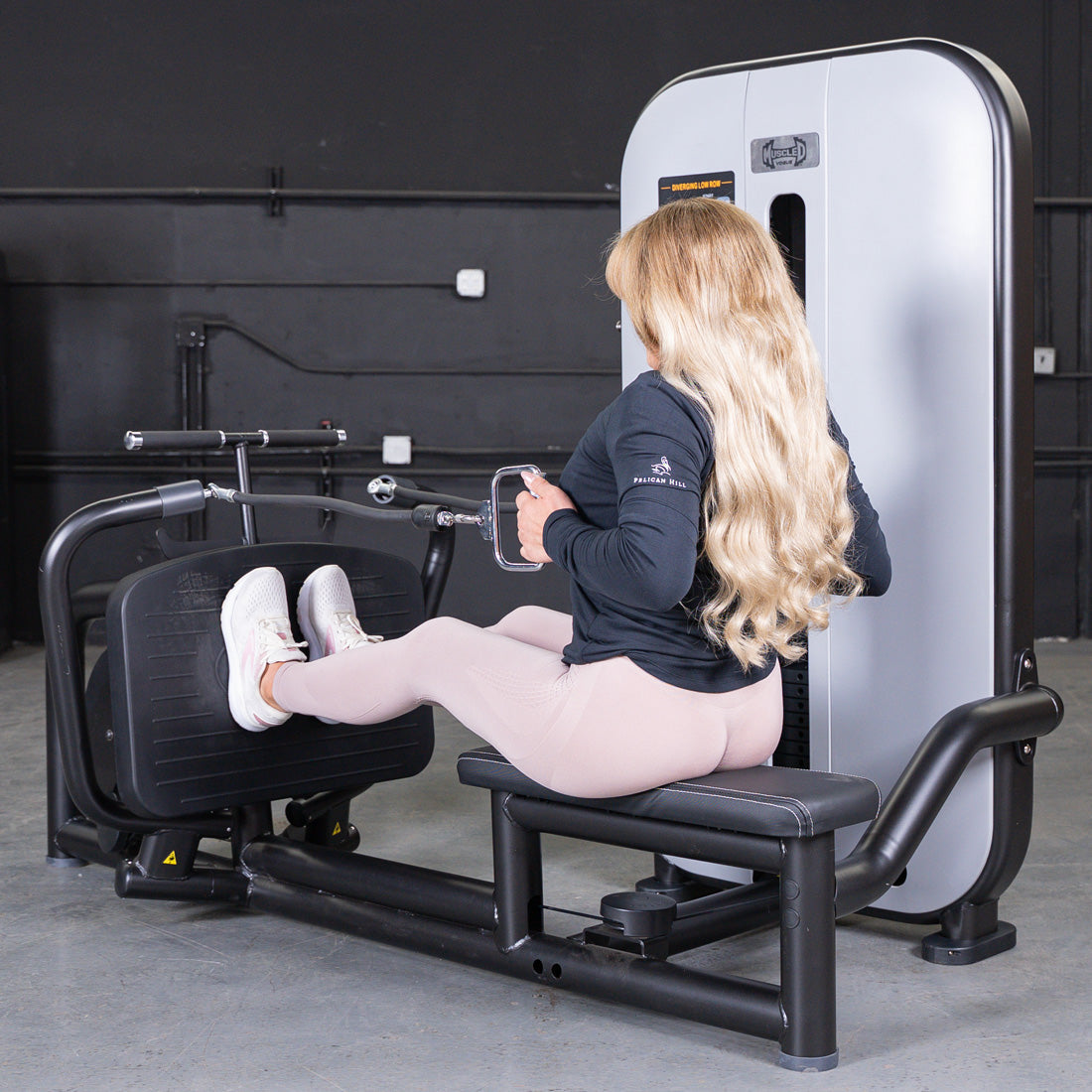 Woman with long blonde hair using a seated low row machine with feet on footrests and hands on handles.