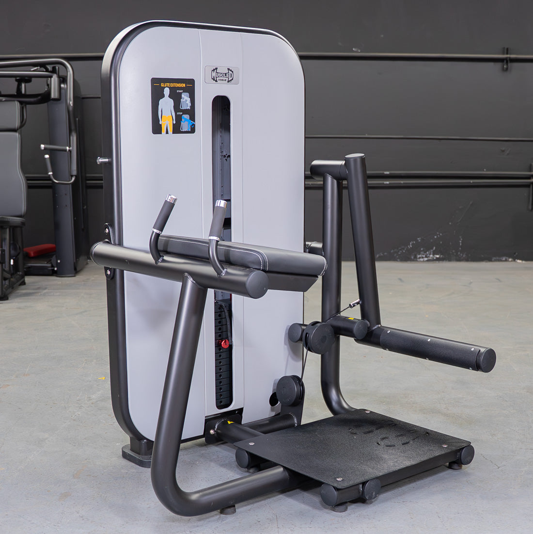 Gray and black glute extension machine with a weight stack, padded armrests, and foot platform.