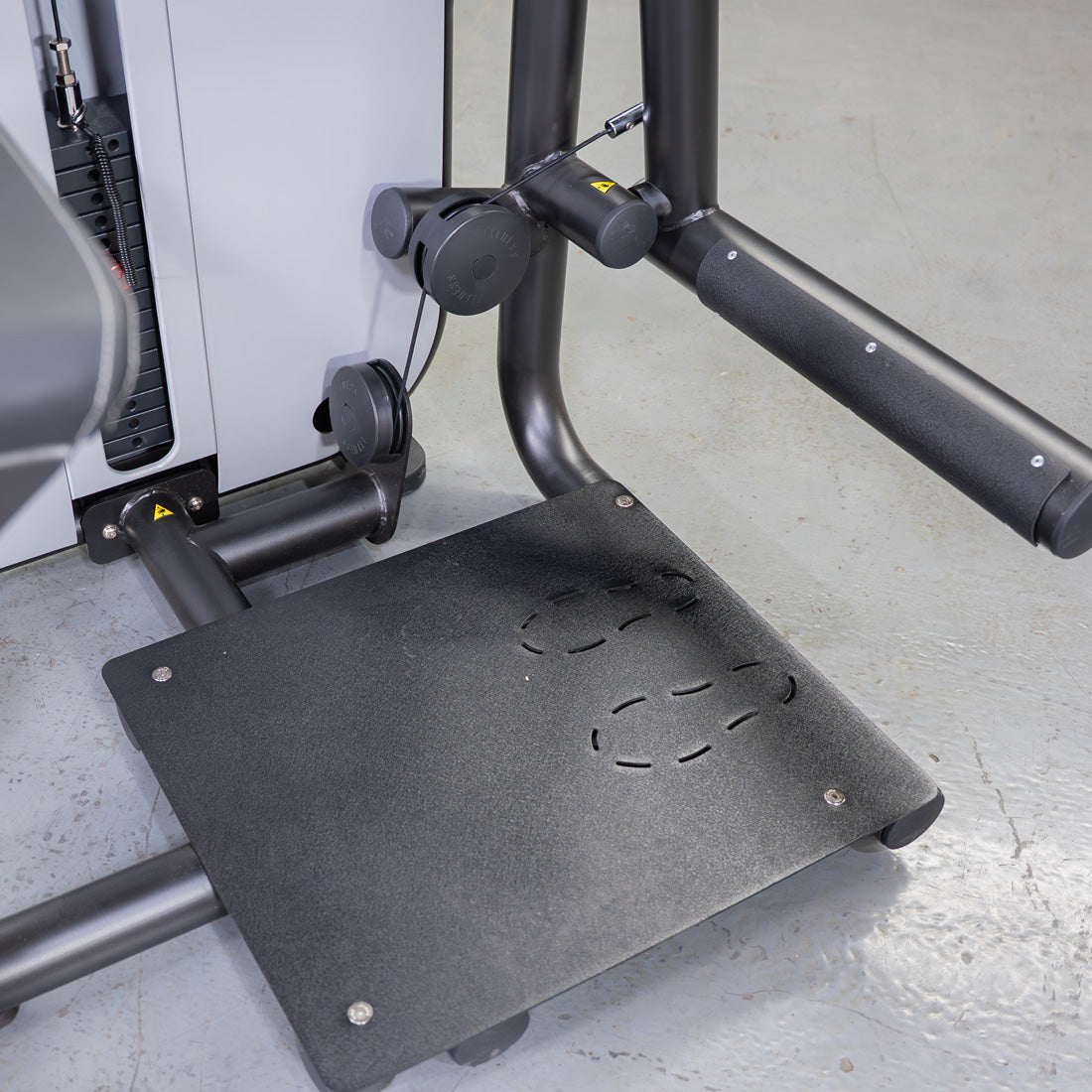 Close-up of a black glute extension machine with a textured footplate and padded armrest.