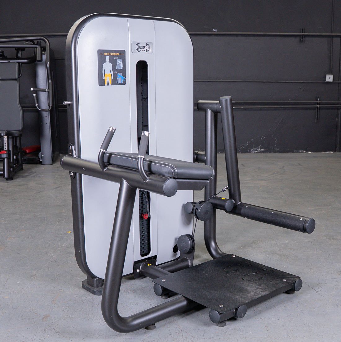 Gray and black glute extension machine with a padded armrest and foot platform in a gym setting.