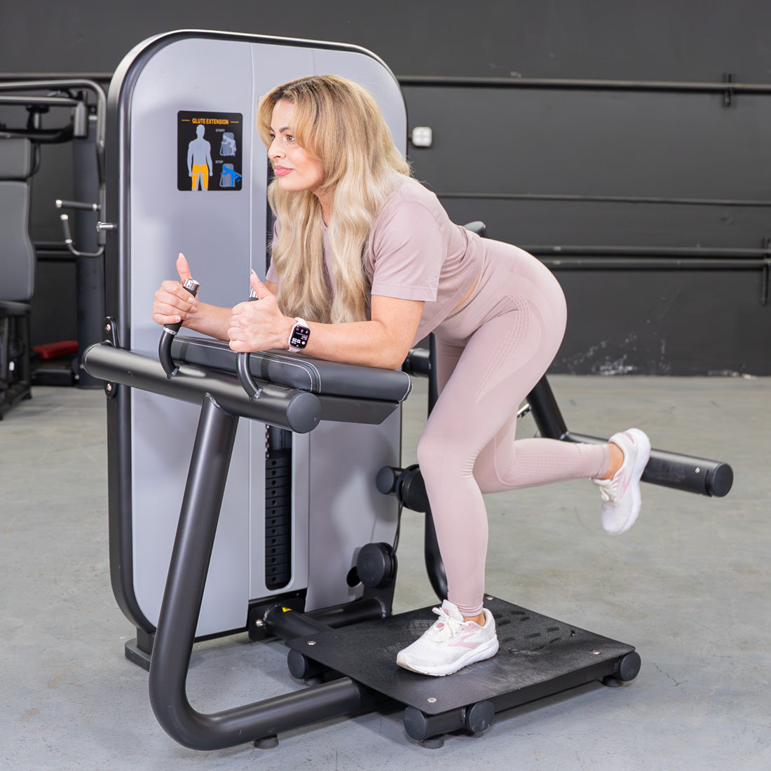Woman in pink activewear using a glute extension machine, giving thumbs up.