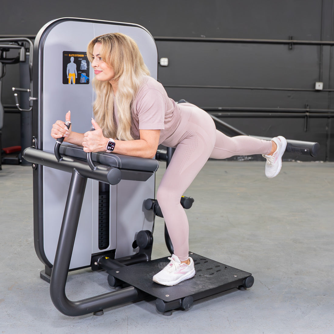 Woman in pink activewear using a glute extension machine, giving two thumbs up.