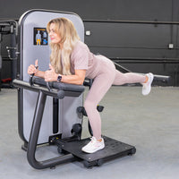 Thumbnail for Woman in pink activewear using a glute extension machine, giving two thumbs up.