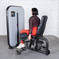 Thumbnail for Woman using a black and gray inner/outer thigh workout machine with red leggings and a black sheer top.
