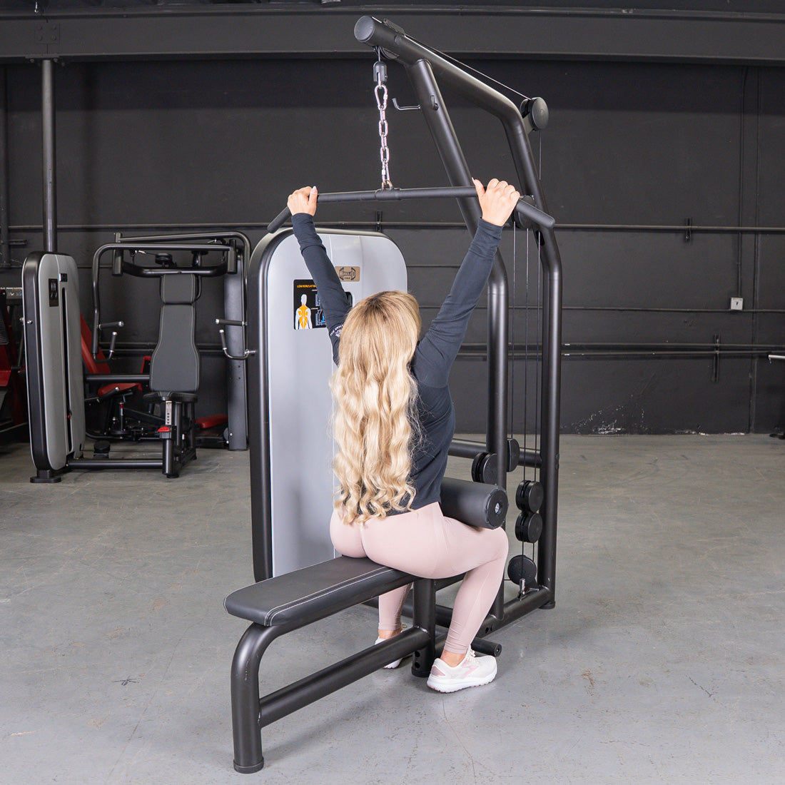 Woman with long blonde hair using a lat pulldown machine in a gym.