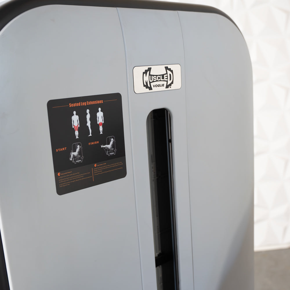 Close-up of a gray leg extension machine with a "Muscle D Vogue" logo and a "Seated Leg Extensions" instruction sticker.