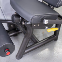 Thumbnail for Close-up of a leg curl machine's black padded seat, yellow adjustment handle, and leg roller with a red button.