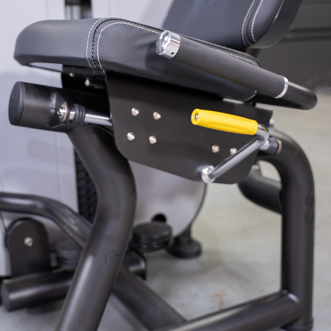 Close-up of a black seated leg curl machine with a yellow adjustment handle and white stitching on the seat.