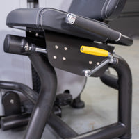 Thumbnail for Close-up of a black seated leg curl machine with a yellow adjustment handle and white stitching on the seat.