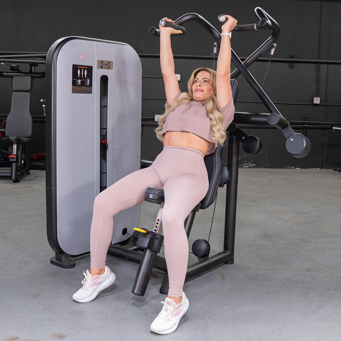 Woman in pink activewear using a shoulder press machine in a gym.