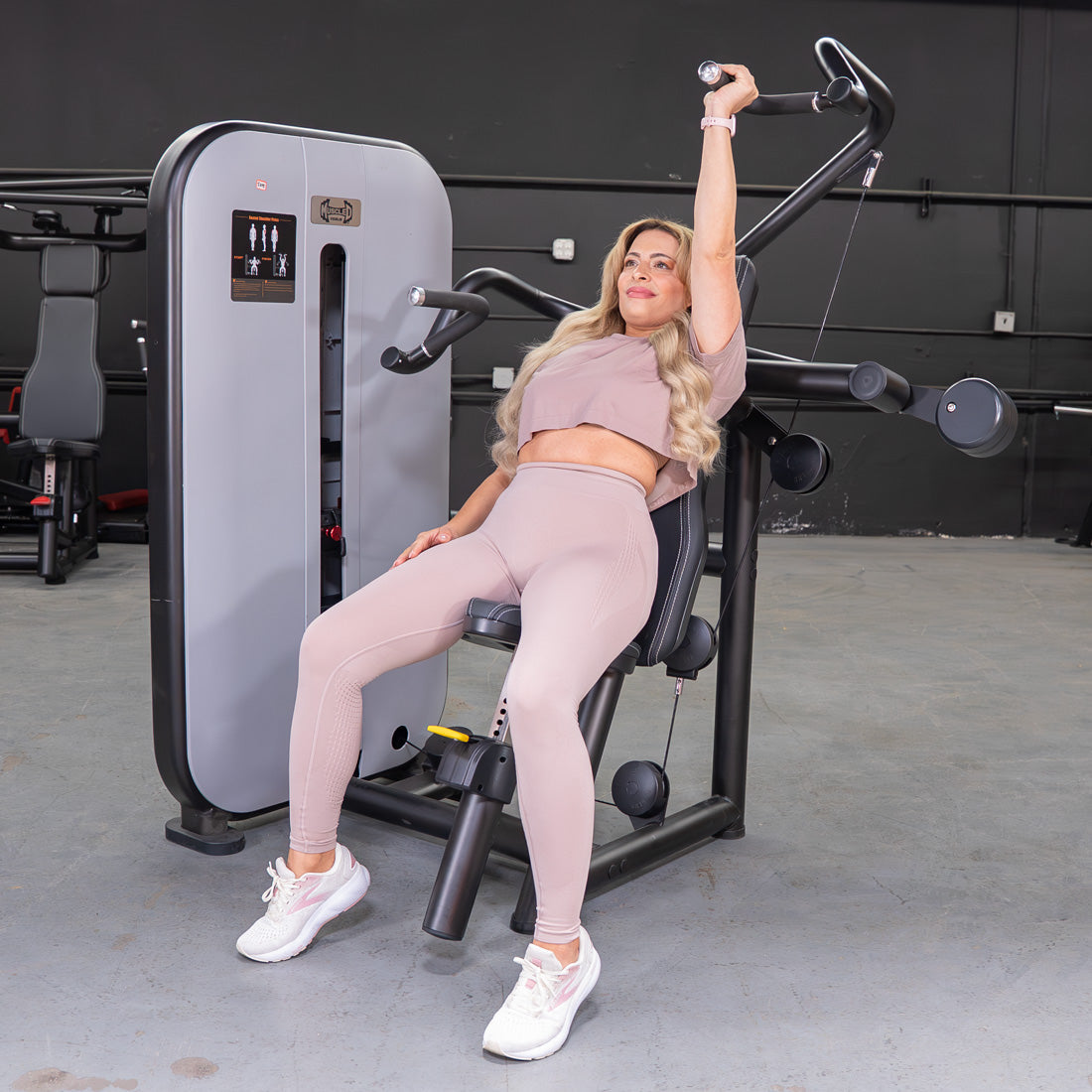 Woman in activewear using a shoulder press machine with one arm extended upwards.