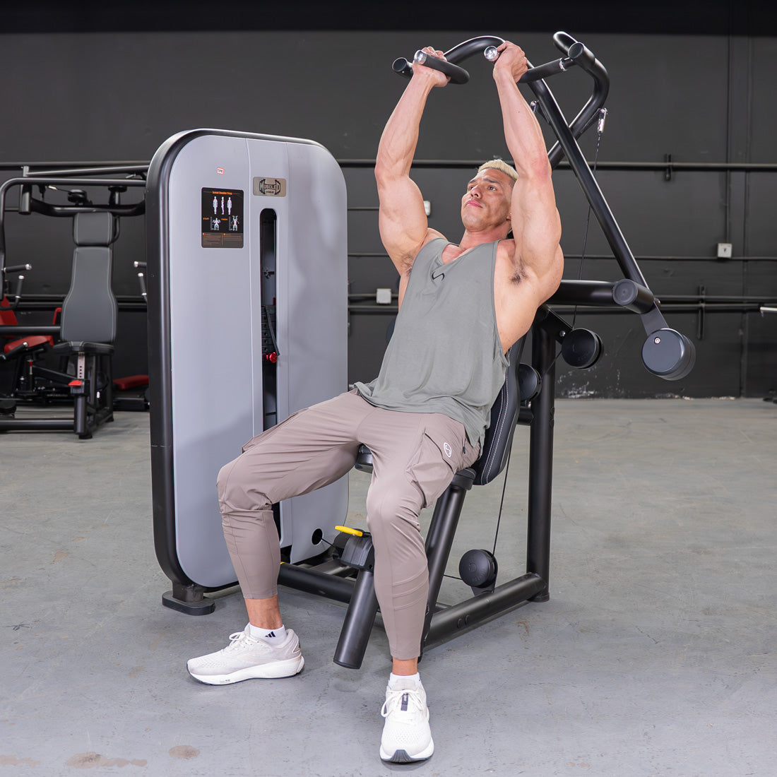 Man using a black and gray shoulder press machine with an adjustable seat in a gym.