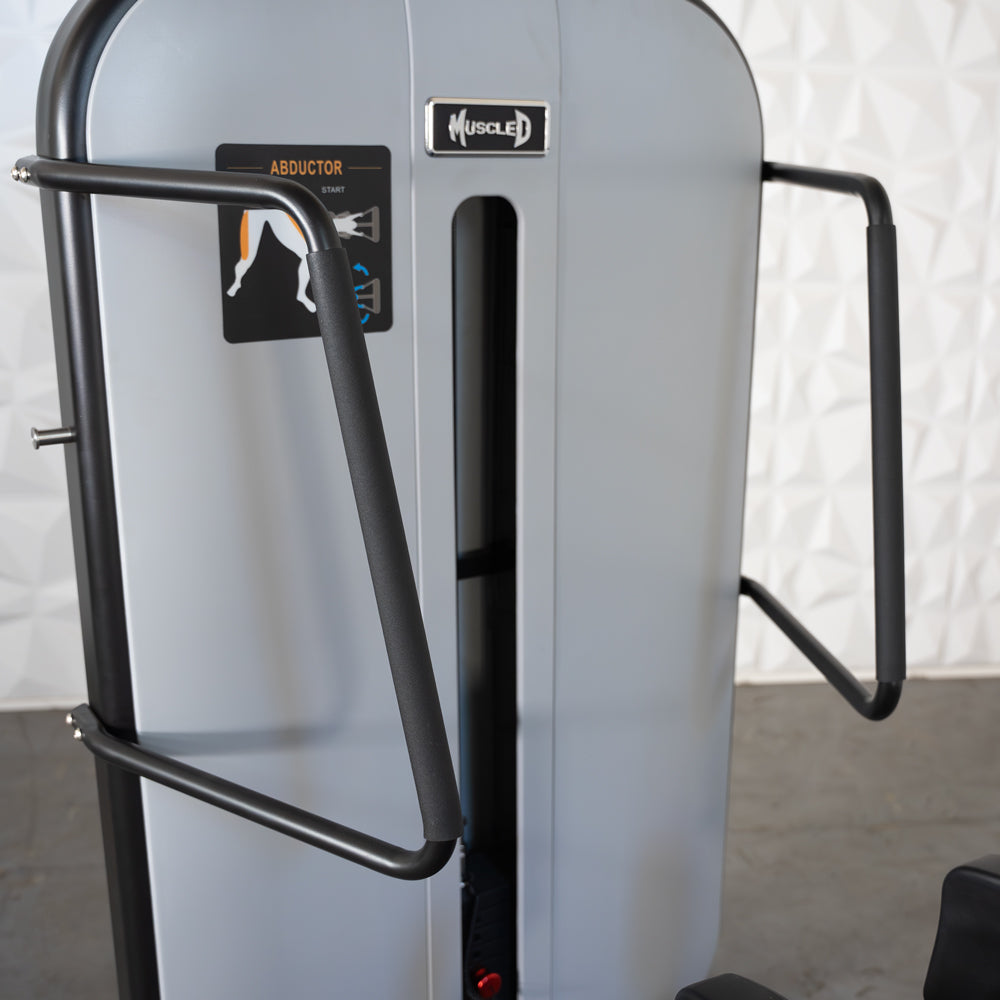 Close-up of a gray standing abductor machine with black padded handles and a "Muscle D" logo.