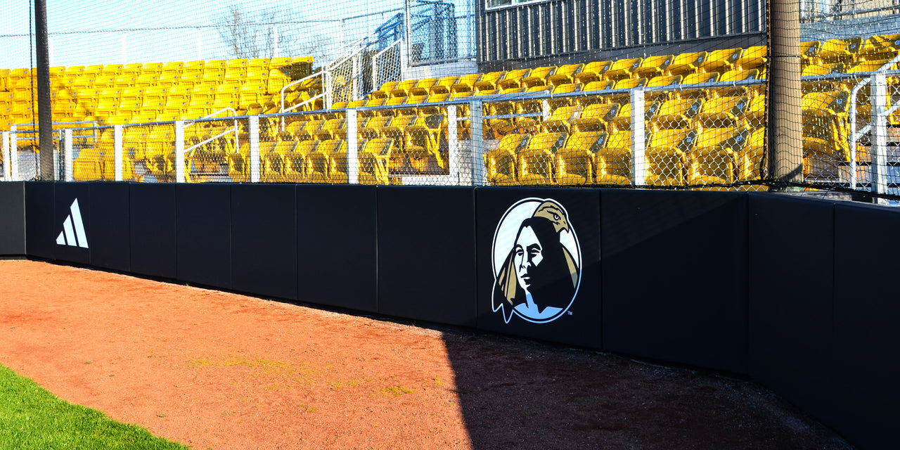 Black backstop wall padding with a school logo in front of yellow stadium seats