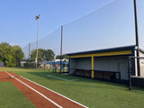 Tall black backstop netting suspended from poles along a turf baseball field's foul line, with a covered dugout to the right.