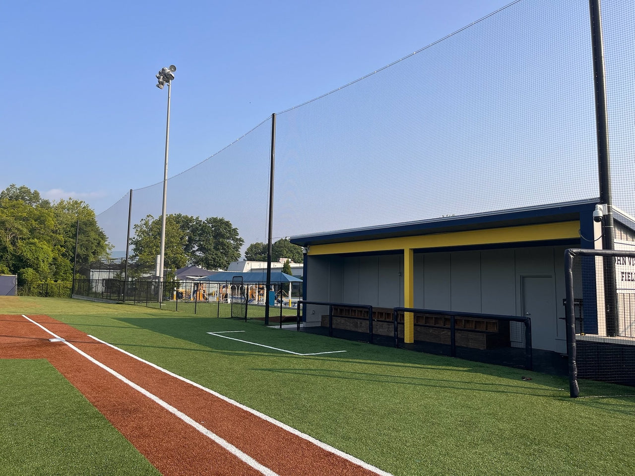 Tall black backstop netting suspended from poles along a turf baseball field's foul line, with a covered dugout to the right.