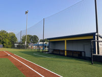 Thumbnail for Tall black backstop netting suspended from poles along a turf baseball field's foul line, with a covered dugout to the right.