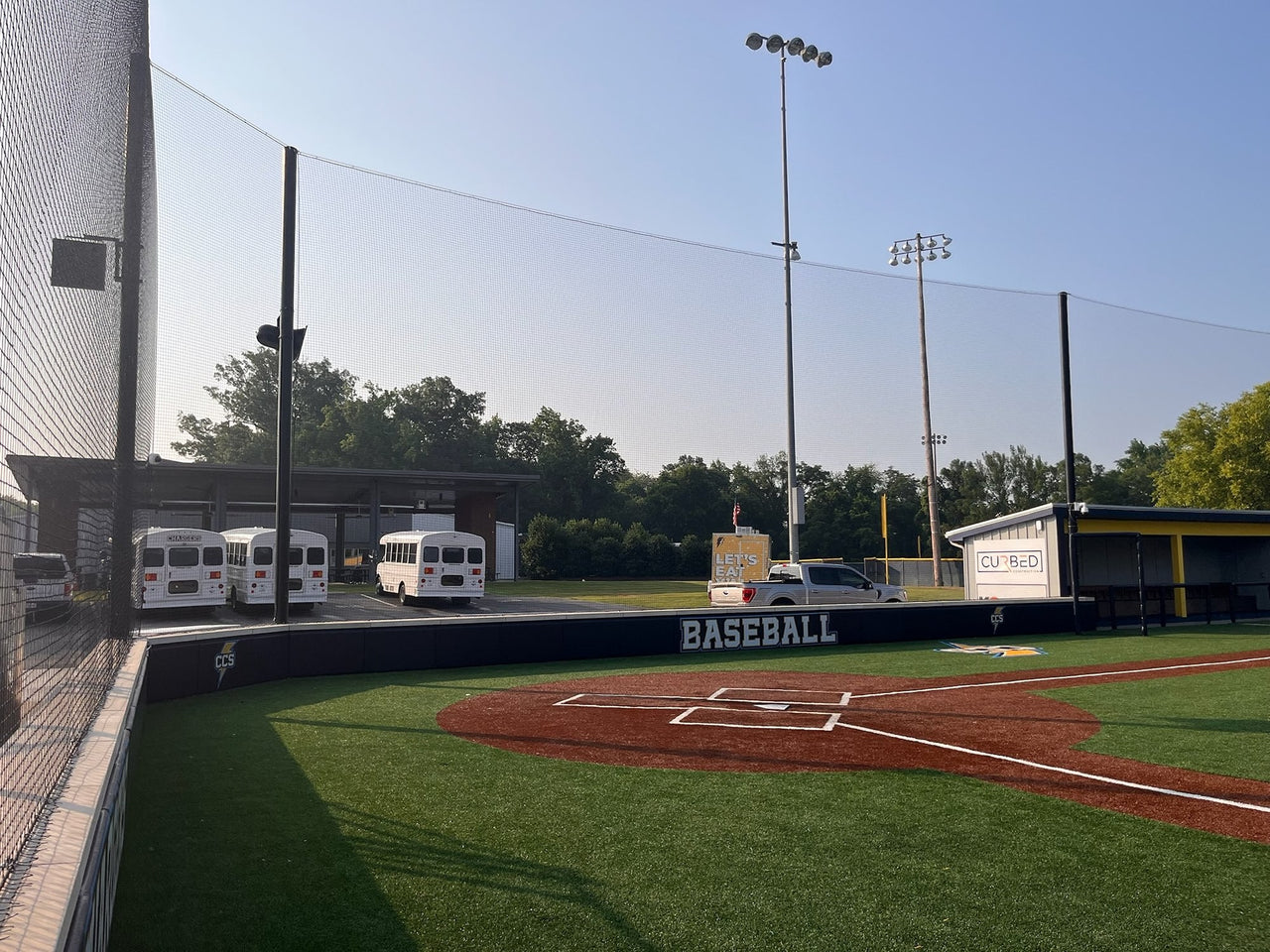 Tall baseball backstop netting curving behind a padded home-plate wall marked Baseball and CCS, with Chargers team buses and a covered dugout beyond.