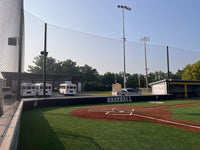 Thumbnail for Tall baseball backstop netting curving behind a padded home-plate wall marked Baseball and CCS, with Chargers team buses and a covered dugout beyond.