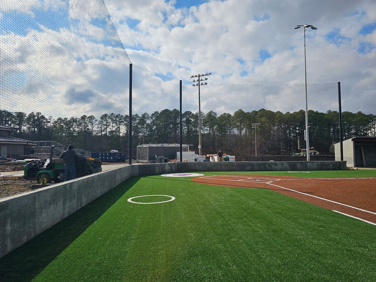 Tall black backstop netting strung between poles behind the home-plate wall of a turf baseball field, with on-deck circles and a utility cart at left.